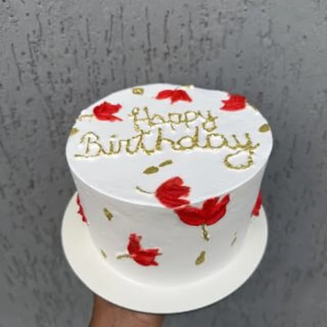 Bolo branco com flores vermelhas pintadas à mão e escrita 'Happy Birthday' em dourado